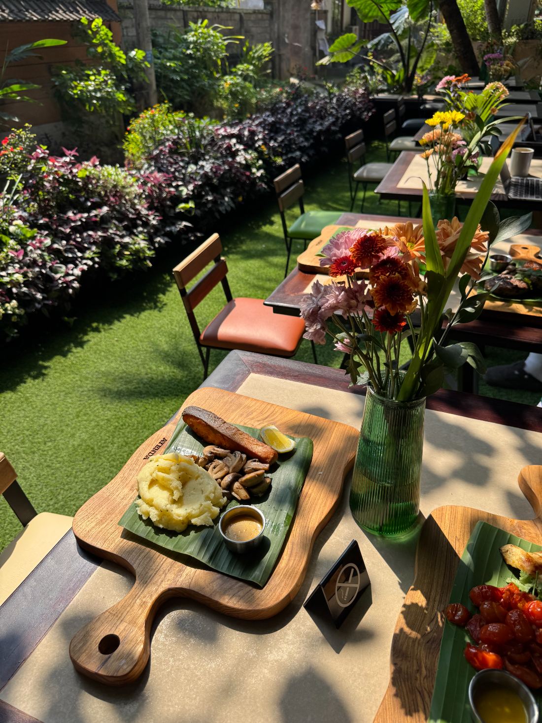 A grilled fillet on a banana leaf and a wooden board, on a café table beside the flower beds